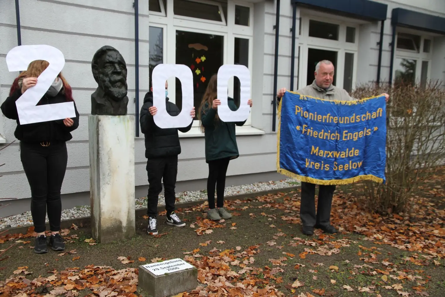 Feierlich: Passend zur Enthüllung der Friedrich-Engels-Geschichtspfad-Tafel an der Neuhardenberger Grundschule, von Schülern gehalten die 200er-Zahl wegen des bevorstehenden Geburtstags, präsentierte Heimatvereinschef-Dietmar Zimmermann auch noch eine alte Pionierfahne.
