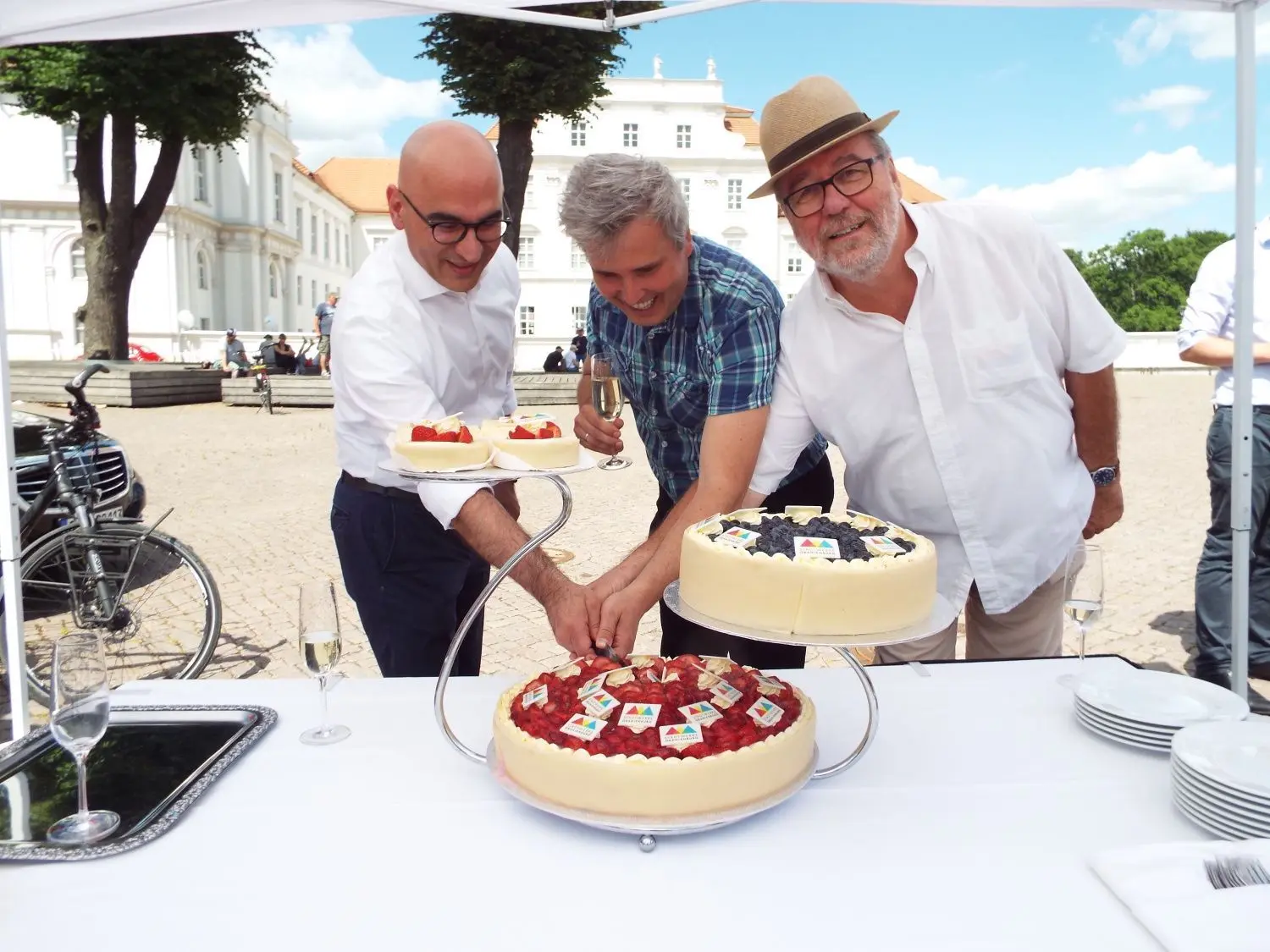 Alireza Assadi, Geschäftsführer der Stadtwerke Oranienburg, der amtierende Bürgermeister der Stadt, Alexander Laesicke (parteilos), sowie Ex-Bürgermeister, Hans-Joachim Laesicke, (von links) haben am Sonntag auf dem städtischen Schlossplatz Erdbeer-, Blaubeer- und Kokostorte angeschnitten sowie verteilt.
