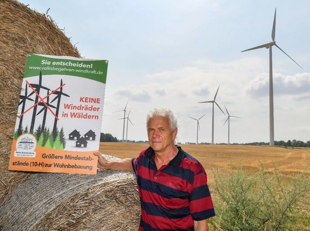 Vor der Haustür: Rainer Ebeling, Sprecher der Bürgerinitiative in Crussow, steht mit einem Protestplakat gegen Windenergie auf einem Feld mit Windrädern.