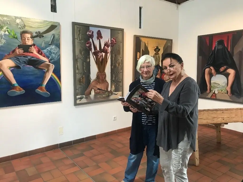 Ein Junge mit Tablet-PC, eine Frau mit Burka: Malerei von Claudia Hauptmann ist zurzeit der Alten Schulscheune zu sehen. Brigitte Fricke (links) und Christine Hannemann vom Förderverein Diensdorf-Radlow blättern in einem Katalog der Künstlerin.