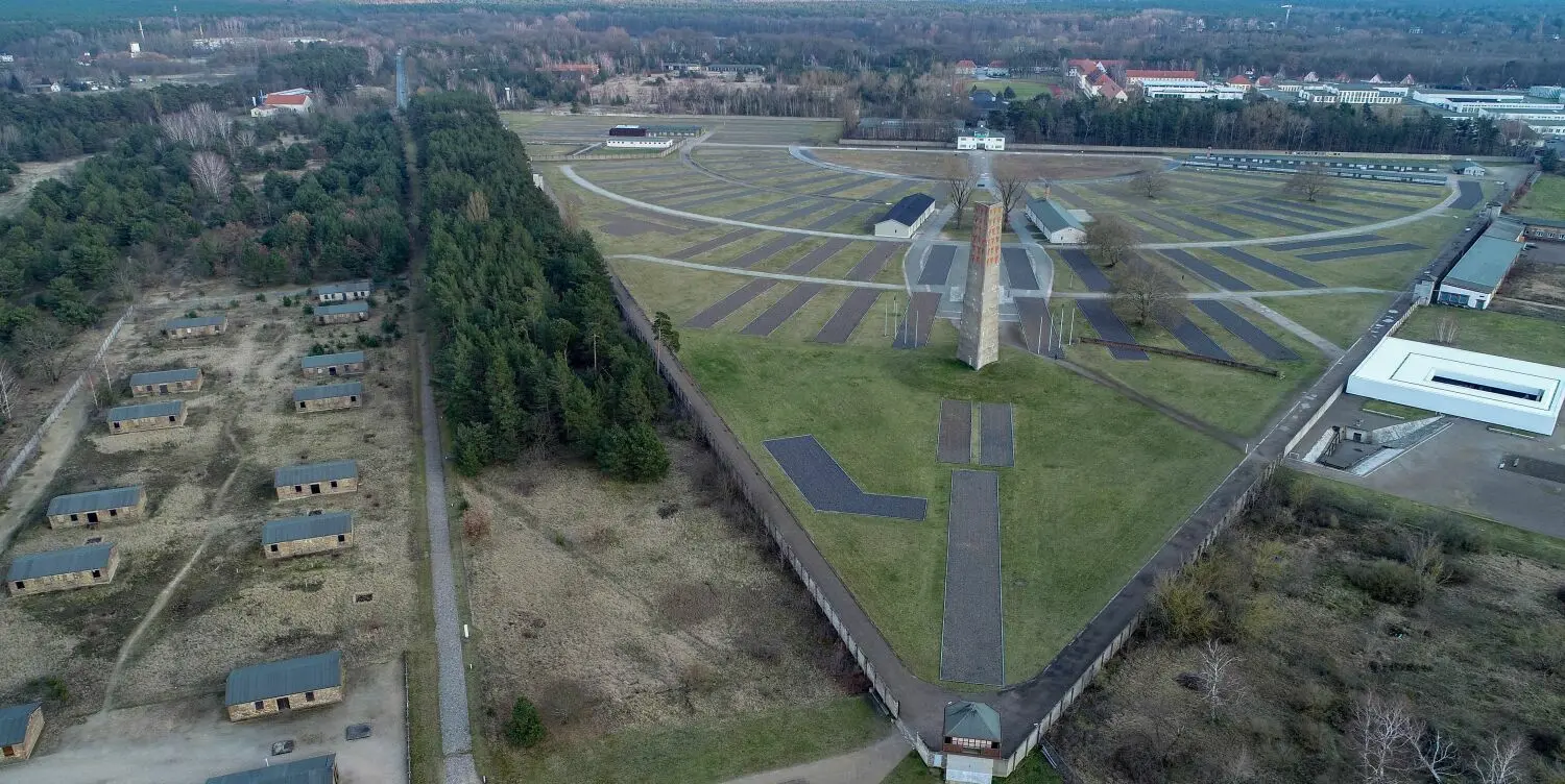 Baracken (l) des KZ-Sonderlagers und Zone II des Speziallagers in der Gedenkstätte Sachsenhausen (Luftaufnahme mit einer Drohne). Etwas rechts im Bild ist das Mahnmal der „Nationalen Mahn- und Gedenkstätte“ aus dem Jahr 1961 zu sehen. Der etwa 40 Meter hohe Obelisk war das zentrale Mahnmal und das Wahrzeichen der 1961 eröffneten Nationalen Mahn- und Gedenkstätte Sachsenhausen der DDR. Die Unterkunftsbaracken wurden in vier Reihen um den halbkreisförmigen Appellplatz errichtet. Das KZ Sachsenhausen wurde im Sommer 1936 von Häftlingen errichtet.