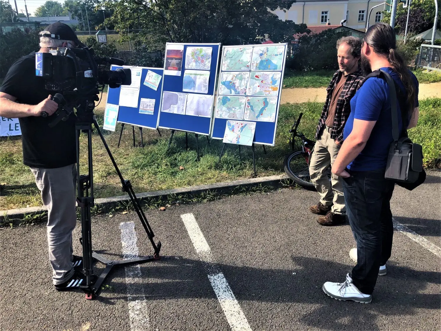 Vor der Stadthalle am Tag der Erörterungen zur Tesla-Baustelle: Infowand der BI Grünheide mit Infomaterial und Karten zu geplanten Veränderungen in der Region. Kameramann vom RBB (v.l.), Vertreter der BI Grünheide und Tobias Lindh aus Erkner, der den Bau des Teslawerks in Grünheide begrüßt.