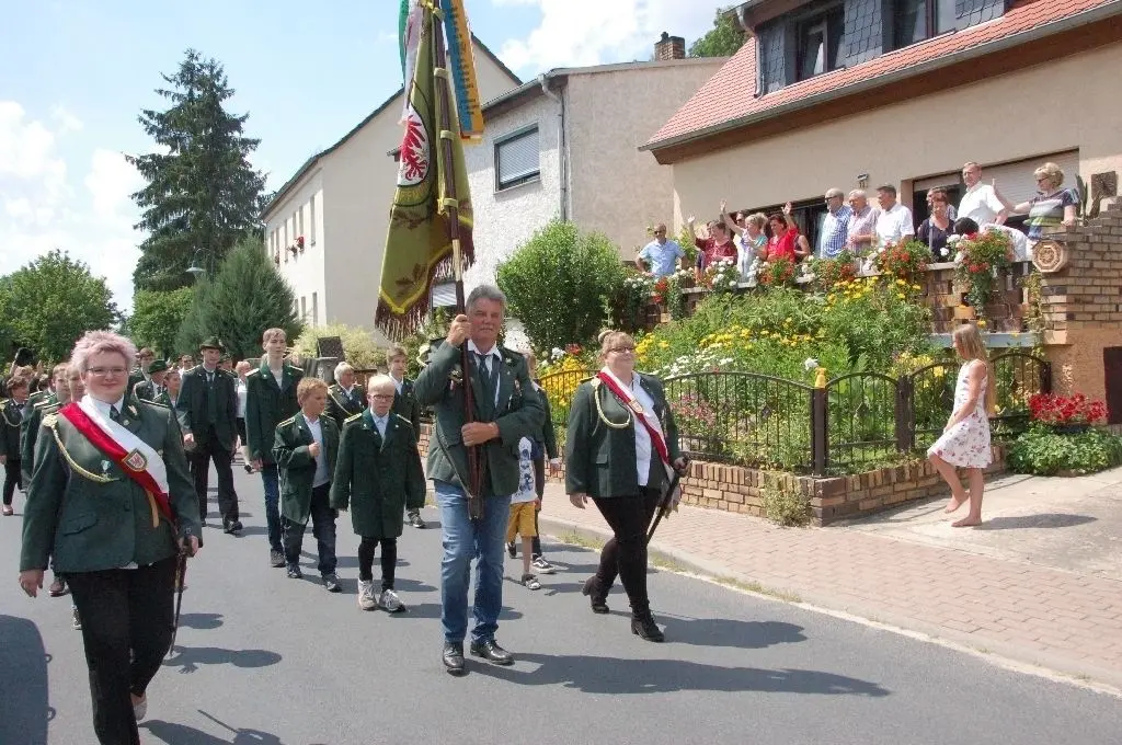 Umzug: Mitglieder des Schützenvereins Schiffmühle marschieren als Gastgeber an der Spitze hinter dem Schulzendorfer Blasorchester. von einem Balkon winken ihnen Zuschauer zu.