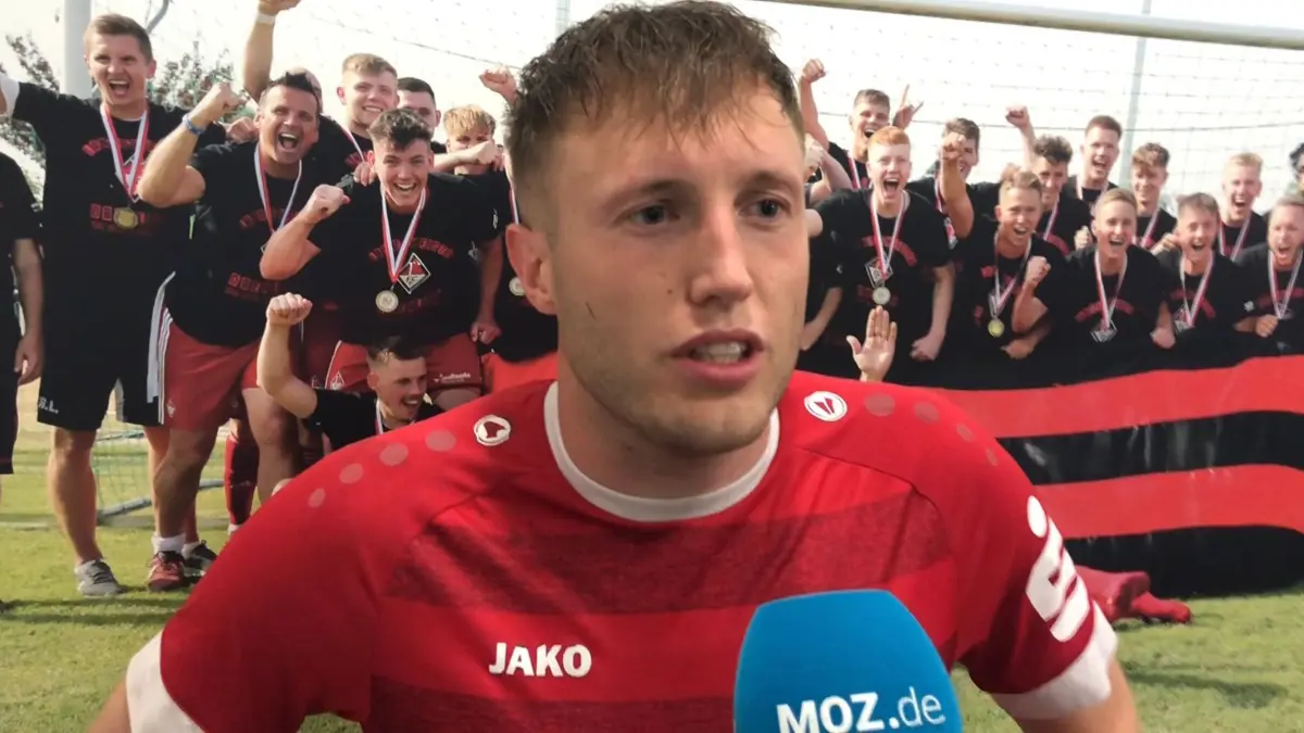 Lukas Sauer vom 1. FC Frankfurt spricht am MOZ-Mikrofon über den guten Saisonstart in der Fußball-Brandenburgliga. Hinter ihm im Stadion der Freundschaft hängt ein Bild von der Meisterfeier im Juni 2022, als dem Verein der Aufstieg in die Oberliga gelang.