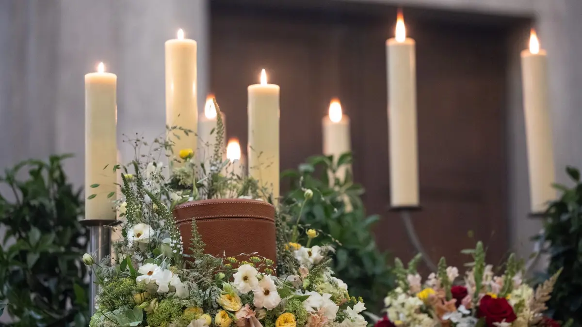 Symbolbild - Beerdigung: PRODUKTION - 12.04.2024, Hessen, Frankfurt/Main: Eine Urne steht umgeben von brennenden Kerzen und Blumen auf einem Podest in der Trauerhalle des Frankfurter Hauptfriedhofs. (zu dpa: «Verband: Mehr Menschen wollen eigene Beerdigung mitbestimmen») Foto: Hannes P Albert/dpa +++ dpa-Bildfunk +++