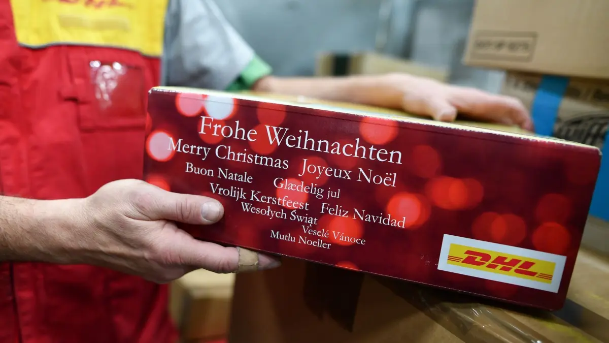 Weihnachtspaket: ARCHIV - 01.12.2016, Rheinland-Pfalz, Speyer: Ein Mitarbeiter der Deutschen Post DHL hält in der Zollabfertigungsstelle des Paketzentrums der Deutschen Post Feldpost mit der Aufschrift «Frohe Weihnachten», die in verschiedenen Sprachen aufgeführt ist, in den Händen. (zu dpa: «Fallstricke im Weihnachtshandel - davor warnt der Zoll») Foto: Uwe Anspach/dpa +++ dpa-Bildfunk +++