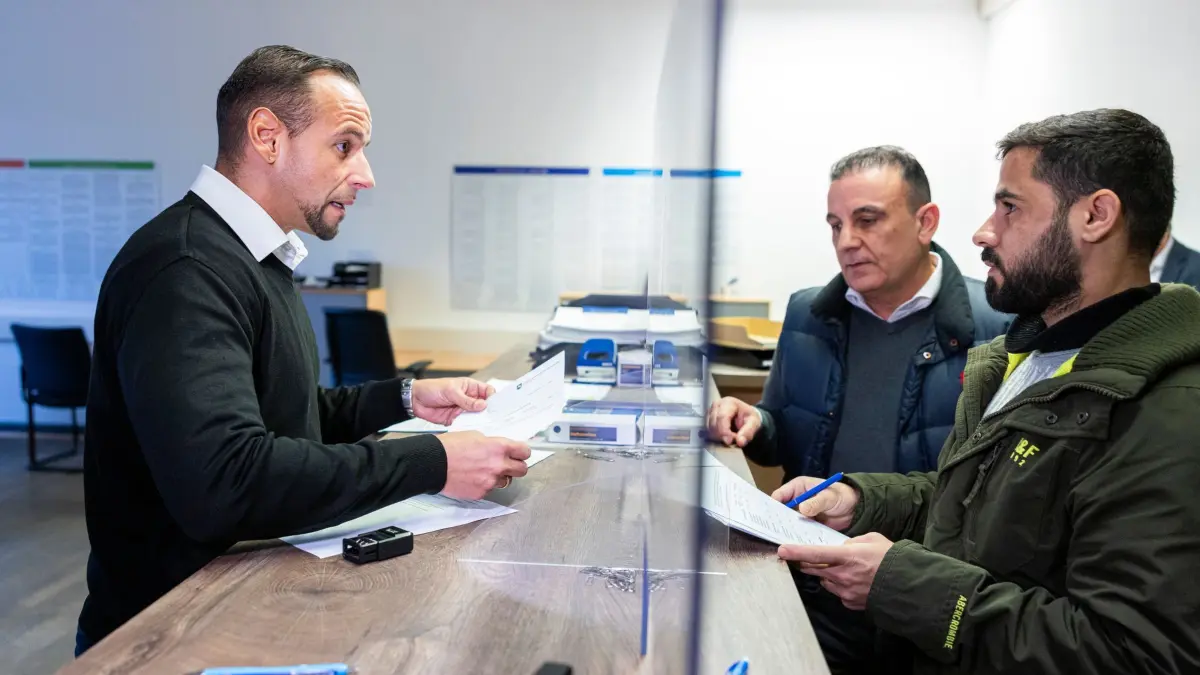 Ausgabe der Bezahlkarte für Geflüchtete in Niedersachsen: 16.12.2024, Niedersachsen, Braunschweig: Semir von Lewinski (l), Mitarbeiter der Landesaufnahmebehörde Niedersachsen, steht während der Ausgabe der Bezahlkarte an Mohammad (r), Geflüchteter aus Syrien, hinter einem Tresen. Daneben steht ein Dolmetscher (2.v.l). Der Bund hat mit der Änderung des Asylbewerberleistungsgesetzes die Möglichkeit eröffnet, dass auch Geflüchtete mit einer Bezahlkarte ohne Bargeld zahlen können. Zunächst sollen die etwa 4.100 Bewohner der Landaufnahmebehörde mit den Karten versorgt werden. Die Ausgabe in den Kommunen für etwa 28.000 Berechtigte ist ab Februar 2025 vorgesehen. Foto: Michael Matthey/dpa +++ dpa-Bildfunk +++