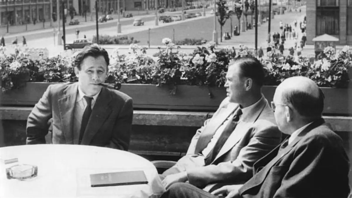 Hermann Henselmann (l.) erläutert am 5.8.1954 auf der Terrasse des Café Warschau in Berlin dem Präsidenten des Nationalrats, Prof. Dr. Correns (r.), und dem ehemaligen Präsidenten des Bonner Verfassungsschutzamtes, Dr. Otto John (Mitte) die Anlage der Stalinallee und die Wiederaufbaupläne des demokratischen Magistrats für die deutsche Hauptstadt. Dr. John besucht in der Stalinallee unter anderem eine Neubauwohnung, das im Bau befindliche Kinderkaufhaus und die Deutsche Sporthalle.