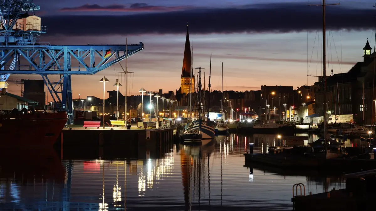 Wetter in Rostock: 28.01.2025, Mecklenburg-Vorpommern, Rostock: Über dem Stadthafen an der Warnow mit der Petrikirche im Hintergrund wird es langsam hell, die Lichter spiegeln sich im glatten Wasser. Foto: Bernd Wüstneck/dpa +++ dpa-Bildfunk +++
