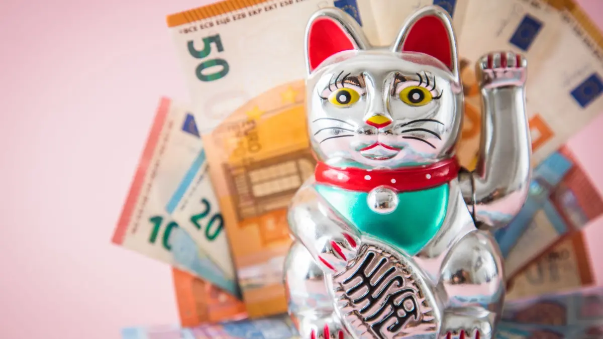 Japanische Winkekatze vor Geldscheinen: ILLUSTRATION - Die japanische Winkekatze soll der Legende nach Glück und Wohlstand herbeiwinken. (zu dpa: «Finanziell stabil? Wie Geldsorgen die Gesundheit belasten») Foto: Christin Klose/dpa-tmn - Honorarfrei nur für Bezieher des dpa-Themendienstes +++ dpa-Themendienst +++