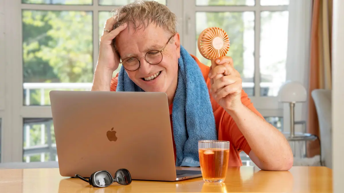 Mann schwitzt im Home Office, leidet unter Hitze in der Wohnung, kühlt sich mit einem Ventilator und einem Getränk, München, Juli 2025: Mann schwitzt im Home Office, leidet unter Hitze in der Wohnung, kühlt sich mit einem Ventilator und einem Getränk, München, Juli 2025 Deutschland, München, Juli 2025, Mann schwitzt im Home Office, leidet unter Hitze in der Wohnung, gönnt sich ein kühles Getränk, Glas Aperol, sitzt am Schreibtisch, kühlt sich mit einem Handventilator, chinesisches Modell aus dem Internet, Schweiß läuft ihm übers Gesicht, er arbeitet am Computer, Alkohol in der Arbeitszeit, Handtuch liegt um die Schultern, schwitzen, Brille, Wohnung, Hitzewelle, Rekordhitze, Sommer, Hochsommer, Bayern, *** Man sweats in the home office, suffers from heat in the apartment, cools himself with a fan and a drink, Munich, July 2025 Germany, Munich, July 2025, Man sweat