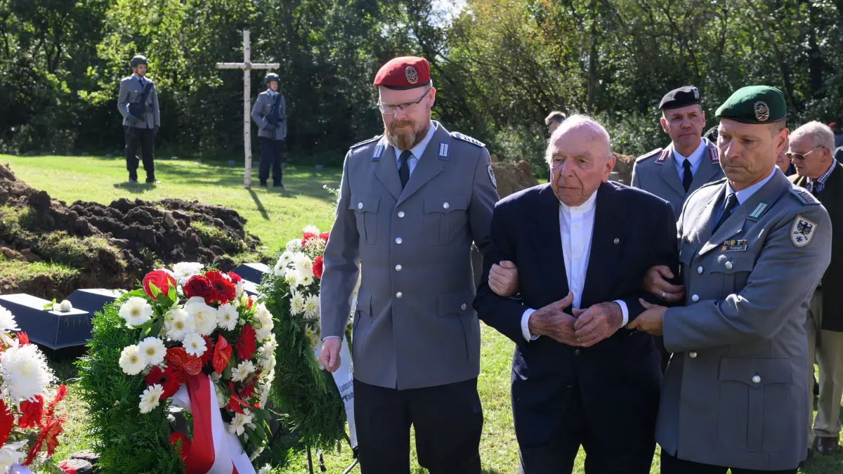 Zweiter Weltkrieg: 60 tote Soldaten in Lietzen eingebettet: 23.09.2025, Brandenburg, Lietzen: Wolf-Dietrich Kroll (M), 98-jähriger Zeitzeuge und ehemaliger Gefreiter vom Fallschirmjäger-Regiment 27 aus dem Zweiten Weltkrieg, wird von Bundeswehrsoldaten auf dem Soldatenfriedhof in Lietzen (Landkreis Märkisch-Oderland) gestützt. Insgesamt 60 im Zweiten Weltkrieg gefallene deutsche Soldaten sind auf der Kriegsgräberstätte zur letzten Ruhe gebettet worden. Die Kriegsgräberstätte in Lietzen wurde bereits während der Kämpfe im Frühjahr 1945 als deutscher Soldatenfriedhof angelegt. Seit 1993 werden dort regelmäßig Gefallene des Zweiten Weltkriegs zur letzten Ruhe gebettet. Foto: Patrick Pleul/dpa +++ dpa-Bildfunk +++