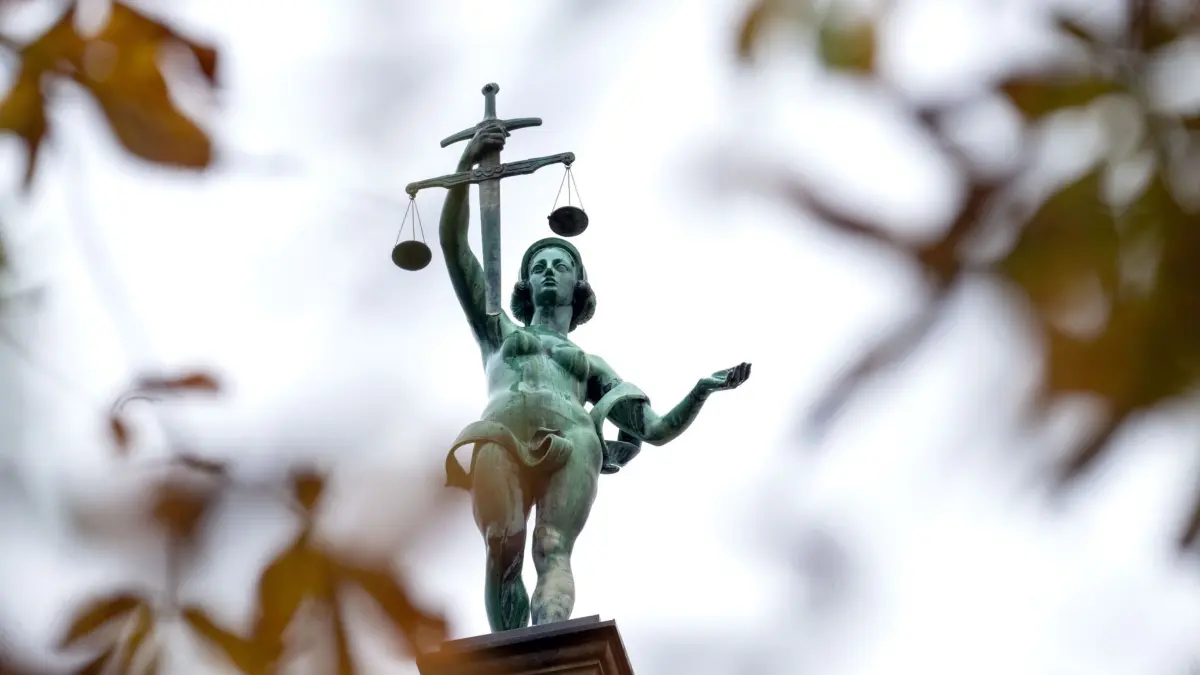 Prozess um Giftanschläge auf Ehefrau beginnt: 23.10.2025, Bayern, Bamberg: Hinter herbstlich verfärbten Blättern steht eine Statue der Justitia auf dem Gebäude des Landgerichts Bamberg. In Bamberg beginnt ein Prozess gegen einen Mann, der mehrfach versucht haben soll, seine Partnerin mit Pflanzengiften zu töten, um mit einer Online-Bekanntschaft ein neues Leben zu beginnen. Die Frau überlebte nur dank mehrerer Klinikaufenthalte und dem Einsetzen eines Herzschrittmachers. Der Angeklagte soll mit seiner neuen Lebensgefährtin nach Frankreich geflüchtet sein, wo er festgenommen wurde. Foto: Pia Bayer/dpa +++ dpa-Bildfunk +++