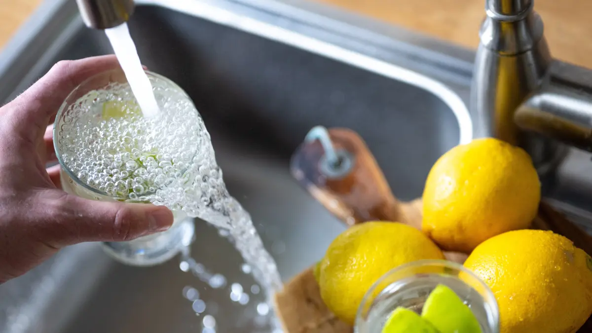 Tinkwasser läuft aus dem Wasserhahn in einer Küche in ein Glas mit Limettenstücken, während im Vordergrund Zitronen auf einem Küchenbrett liegen. (zu dpa: «Bürger in Passau und Umgebung müssen Leitungswasser abkochen») +++ dpa-Bildfunk +++