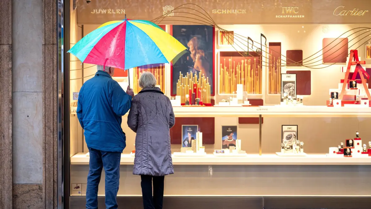 Senioren: 02.12.2025, Bremen: Zwei ältere Personen stehen bei Regen vor einem Schaufenster eines Schmuckgeschäfts und betrachten die ausgestellten Waren. Sie teilen sich einen bunten Regenschirm. Im Bundestag steht am Freitag die Entscheidung über die geplante Rentenreform an, die ein stabiles Rentenniveau, erweiterte Kindererziehungszeiten, steuerfreie Zuverdienstmöglichkeiten im Alter und weitere Änderungen in der Altersvorsorge vorsieht. Foto: Sina Schuldt/dpa +++ dpa-Bildfunk +++