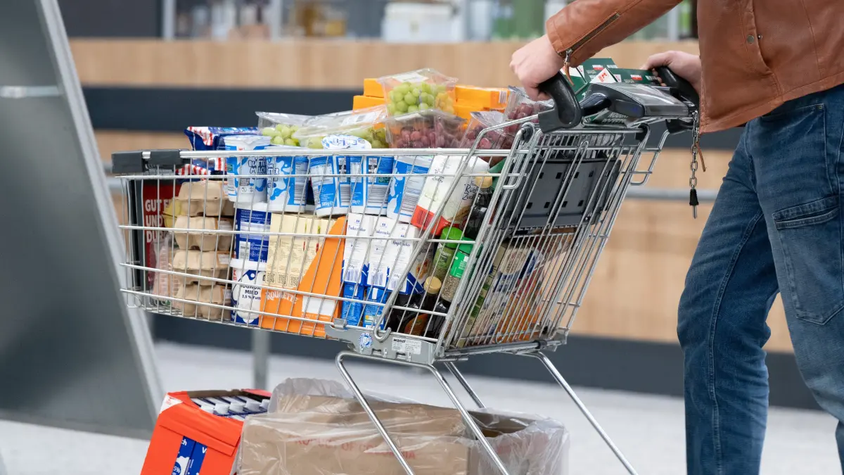 Ein Mann schiebt in einem Supermarkt einen vollen Einkaufswagen mit Lebensmitteln und Getränken.