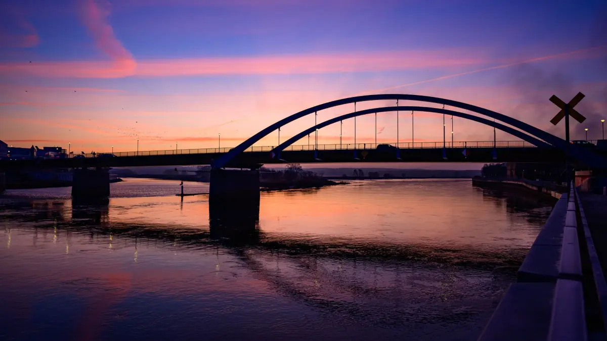 Sonnenaufgang in Brandenburg: 02.12.2025, Brandenburg, Frankfurt (Oder): Der Morgenhimmel leuchtet kurz vor Sonnenaufgang über dem deutsch-polnischen Grenzübergang Stadtbrücke. Mit Wolken und leichtem Frost startet der Tag in Berlin und Brandenburg. Bis zum Vormittag kann es bei null bis minus drei Grad vereinzelt glatt werden, wie der Deutsche Wetterdienst mitteilte. Später steigen die Temperaturen auf maximal drei bis sechs, im äußersten Süden bis sieben Grad. Dabei bleibt es trocken und bedeckt. Foto: Patrick Pleul/dpa +++ dpa-Bildfunk +++