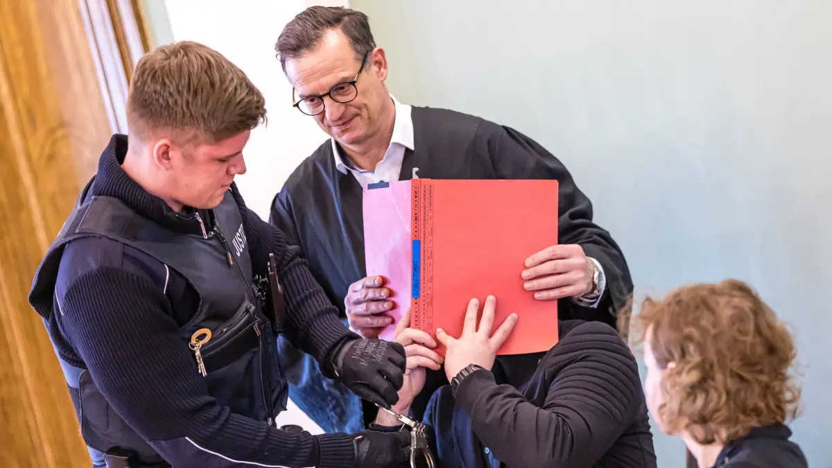 Fortsetzung Prozess nach Tod eines Polizisten in Lauchhammer: 19.01.2026, Brandenburg, Cottbus: Dem Angeklagten (verdeckt) werden im Beisein seines Anwaltes Christoph Rühlmann (hinten) und seiner Dolmetscherin von einem Justizwachtmeister bei der Fortsetzung des Prozesses um den gewaltsamen Tod eines Dresdner Polizisten in Lauchhammer die Handschellen abgenommen. Dem Angeklagten wird Mord an einem 32-jährigen Polizisten vorgeworfen. Bei einem Einsatz gegen mutmaßliche Autodiebe vor einem Jahr in Lauchhammer war dieser von einem Fahrzeug erfasst worden und noch am Einsatzort gestorben. Foto: Frank Hammerschmidt/dpa +++ dpa-Bildfunk +++