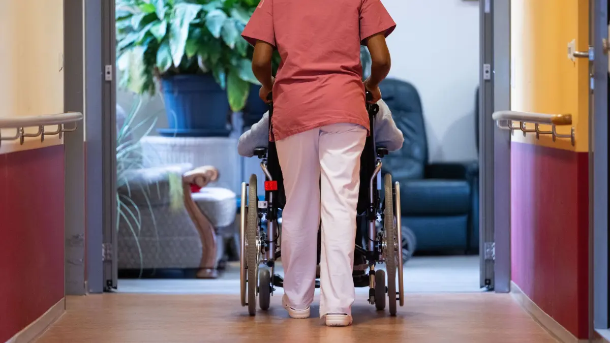 Eine Pflegekraft schiebt einen Pflegebedürftigen im Rollstuhl: ILLUSTRATION - Wohngeld, Pflegewohngeld, Hilfe zur Pflege: Wird das Geld knapp, können Pflegebedürftige im Heim Unterstützung bekommen. (zu dpa: «Geld reicht nicht mehr fürs Pflegeheim - was tun?») Foto: Marijan Murat/dpa/dpa-tmn - Honorarfrei nur für Bezieher des dpa-Themendienstes +++ dpa-Themendienst +++