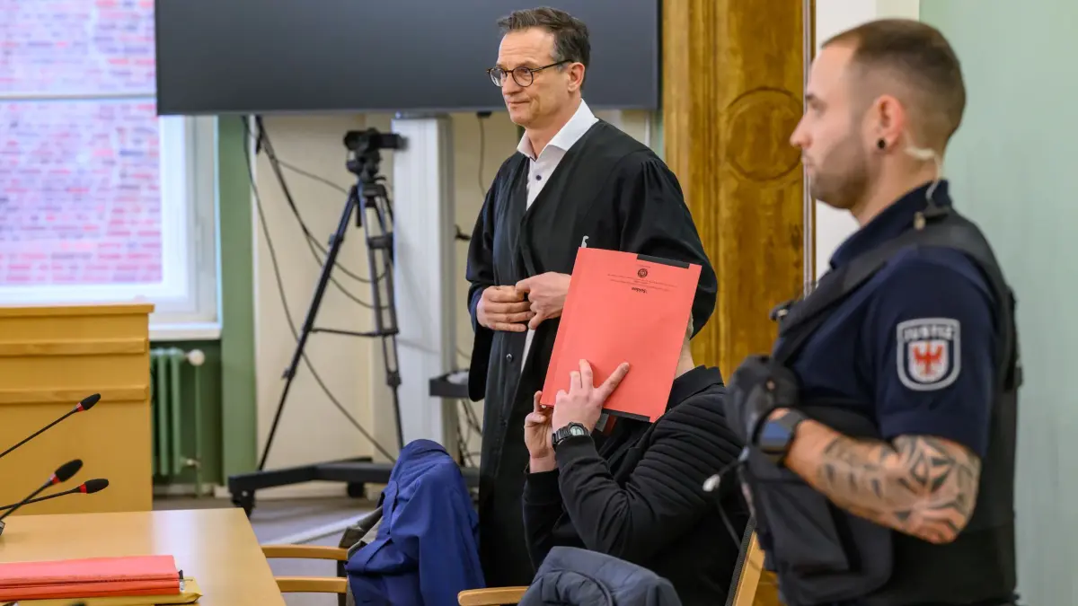 Nach Tod eines Polizisten in Lauchhammer - Prozess beginnt: ARCHIV - 14.01.2026, Brandenburg, Cottbus: In Handschellen und Aktenordner vor dem Gesicht sitzt der Angeklagte im Mordprozess um einen getöteten Polizisten aus Dresden neben seinem Verteidiger Christoph Rühlmann zum Prozessbeginn in einem Verhandlungssaal des Cottbuser Landgerichts. Bei einem Einsatz gegen flüchtende Autodiebe wird ein Polizist aus Dresden in Südbrandenburg von einem Wagen erfasst und getötet. Rund ein Jahr später verhandelt das Landgericht Cottbus wegen des Vorwurfs des Mordes gegen den Fahrer eines gestohlenen Wagens. (zu dpa: «Tod von Polizisten: Angeklagter fuhr wie ein «Rallyefahrer»») Foto: Patrick Pleul/dpa +++ dpa-Bildfunk +++