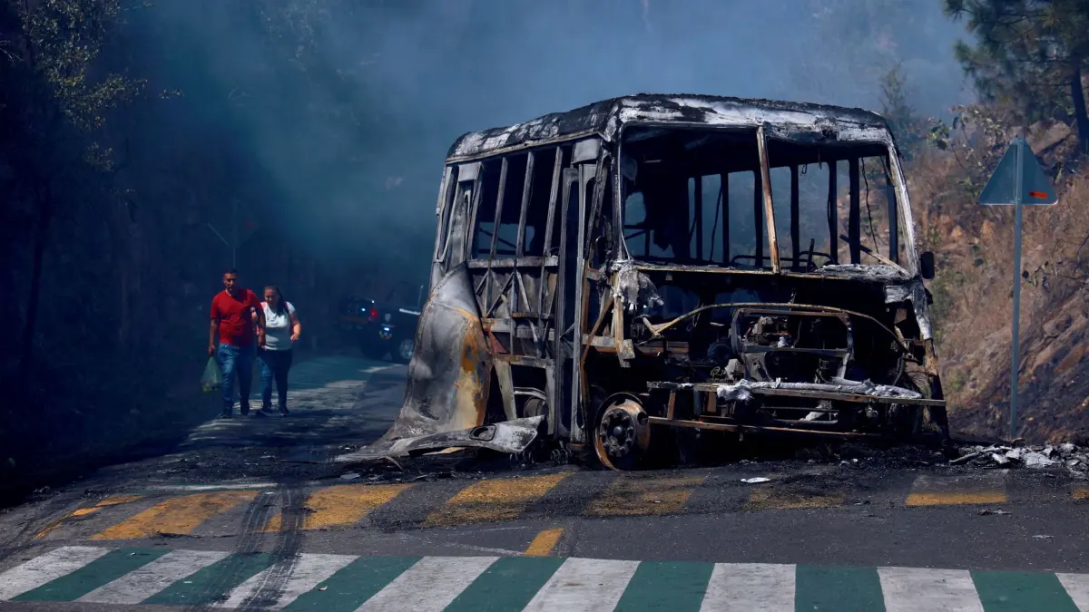 Drogenkrieg in Mexiko - Cointzio: 22.02.2026, Mexiko, Cointzio: Fußgänger gehen an einem ausgebrannten Fahrzeug vorbei, das auf einer Straße in Cointzio im mexikanischen Bundesstaat Michoacán nach dem Tod des Anführers des Kartells der Neuen Generation von Jalisco, «El Mencho», in Brand gesetzt wurde. Foto: Armando Solis/AP/dpa +++ dpa-Bildfunk +++