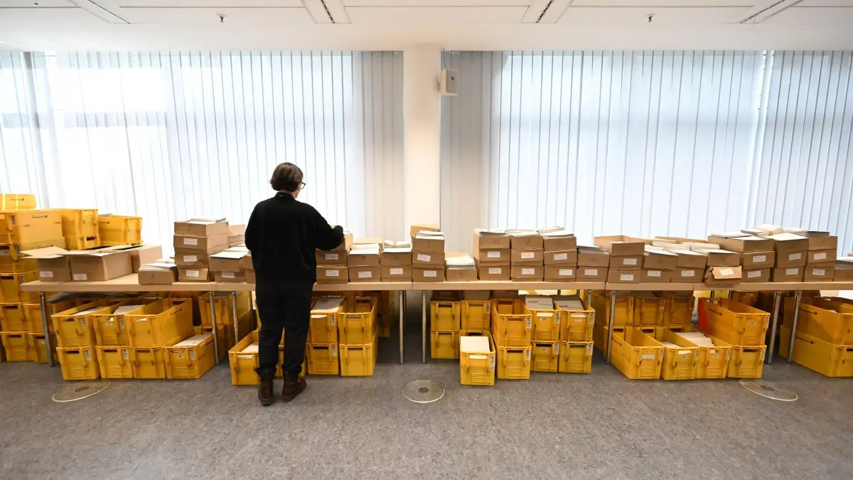 Kommunalwahlen in Bayern: PRODUKTION - 18.02.2026, Bayern, München: Eine Frau steht vor Kisten in denen sich die Wahlunterlagen zur Briefwahl befinden in einer Briefwahlausgabestelle des Münchner Krreisverwaltungsreferat. (zu dpa: «Per Brief oder Urne - Wie wählt Bayern und was sagt uns das?») Foto: Felix Hörhager/dpa +++ dpa-Bildfunk +++