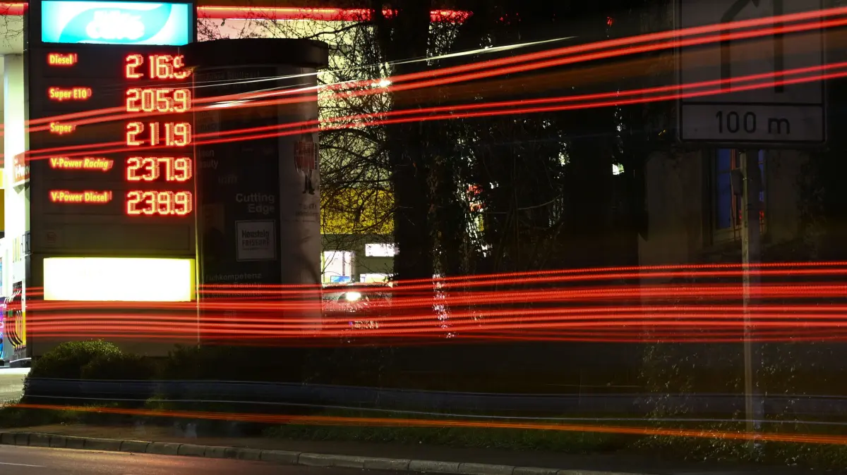 Die Scheinwerfer vorbeifahrender Fahrzeuge ziehen am Morgen auf einer Straße Lichtspuren, während im Hintergrund die Preistafel einer Tankstelle die Kraftstoffpreise anzeigt. (Aufnahme mit langer Belichtungszeit) +++ dpa-Bildfunk +++