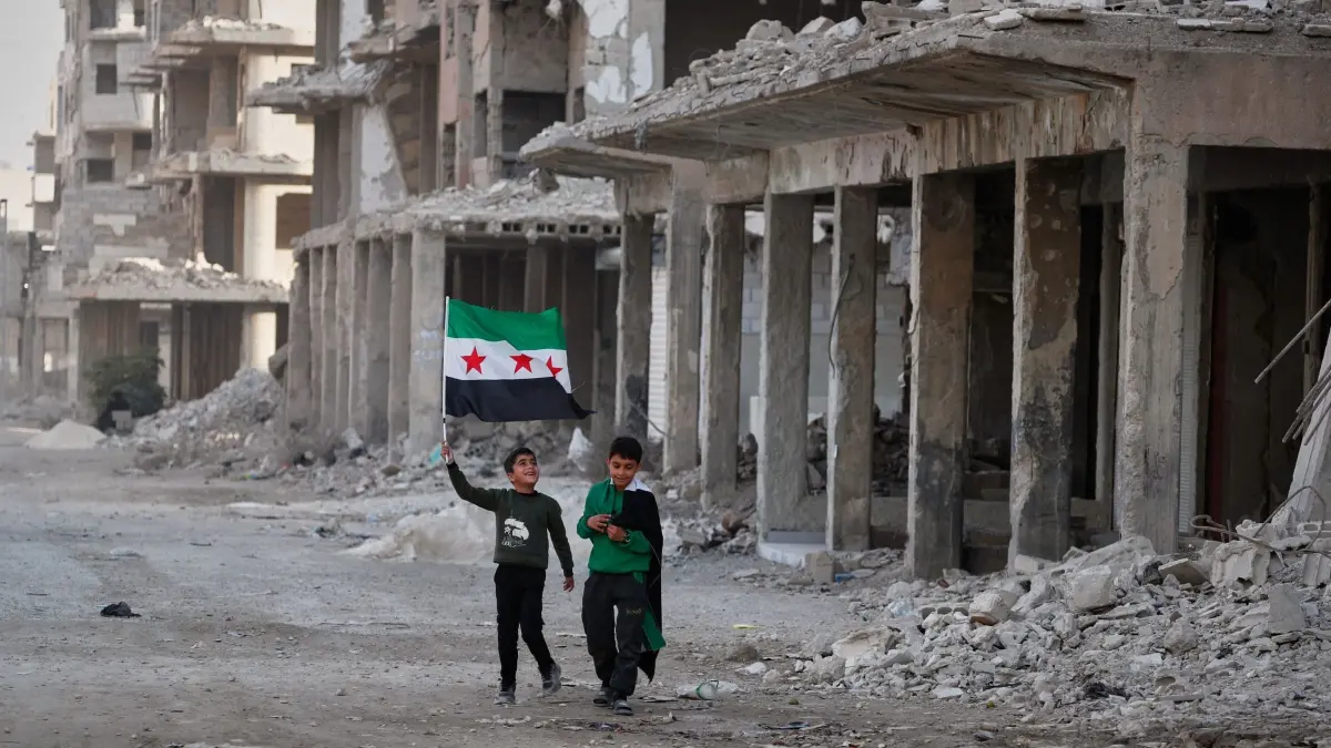 Lage in Syrien - Damaskus: ARCHIV - 05.12.2025, Syrien, Damaskus: Zwei Jungen gehen mit syrischen Flaggen durch eine Straße im kriegszerstörten Damaskus-Vorort Daraya. (zu dpa: ««Beschämend»: Kritik an Merz-Ansage zur Ausreise von Syrern») Foto: Omar Sanadiki/AP/dpa +++ dpa-Bildfunk +++