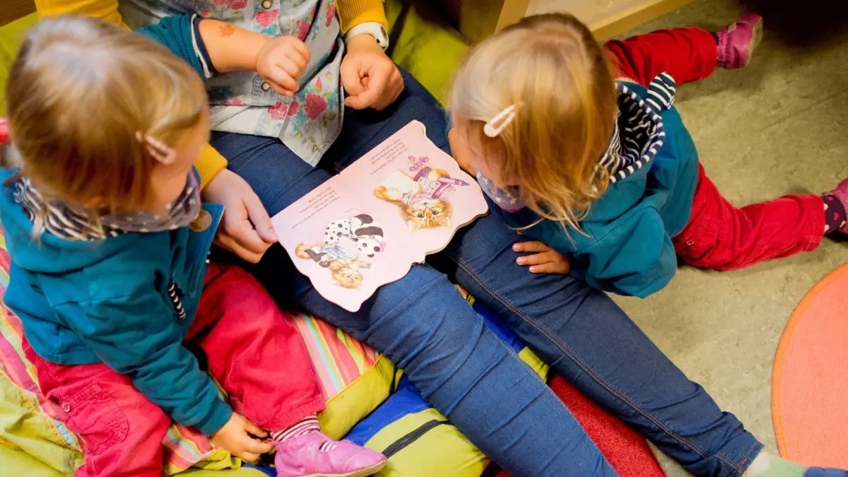 ARCHIV - 16.10.2013, Niedersachsen, Hannover: Eine Erzieherin liest mit Zwillingen ein Buch. Sozialpolitischer Meilenstein in Berlin: Eltern müssen für die Betreuung ihrer Kinder in Kitas oder bei Tagesmüttern künftig generell keine Beiträge mehr zahlen. (zu "Berlin schafft Kita-Gebühren als erstes Bundesland komplett ab" vom 29.07.2018) Foto: Julian Stratenschulte/dpa +++ dpa-Bildfunk +++