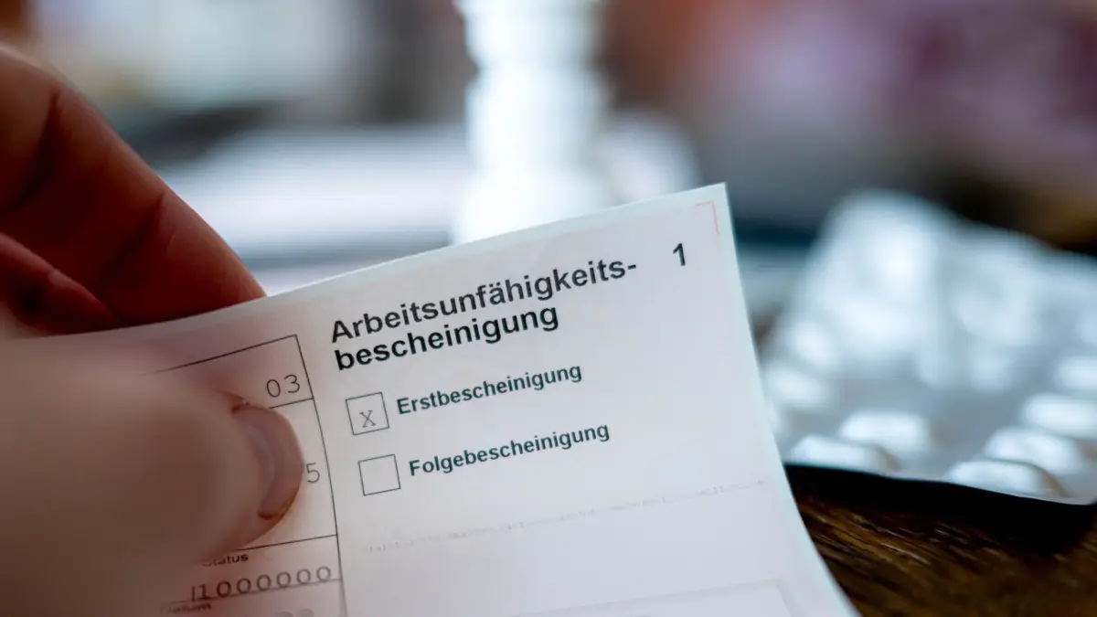 Krankschreibung: ILLUSTRATION - 18.02.2026, Bremen: Eine Person hält eine ausgefüllte Arbeitsunfähigkeitsbescheinigung in der Hand. Im Hintergrund ist unscharf ein Tisch mit Medikamenten zu sehen. (zu dpa: «Kommission empfiehlt Teilkrankschreibung im Job») Foto: Sina Schuldt/dpa +++ dpa-Bildfunk +++