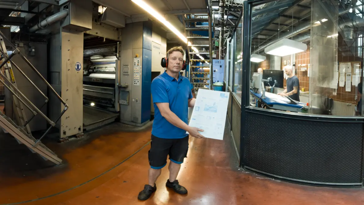 Drucker Jens Zaremba präsentiert eine Druckplatte: Im Hintergrund wird das Zeitungspapier beim Rotationsdruck mit über 30 km/h bedruckt und zu Zeitungen gefaltet