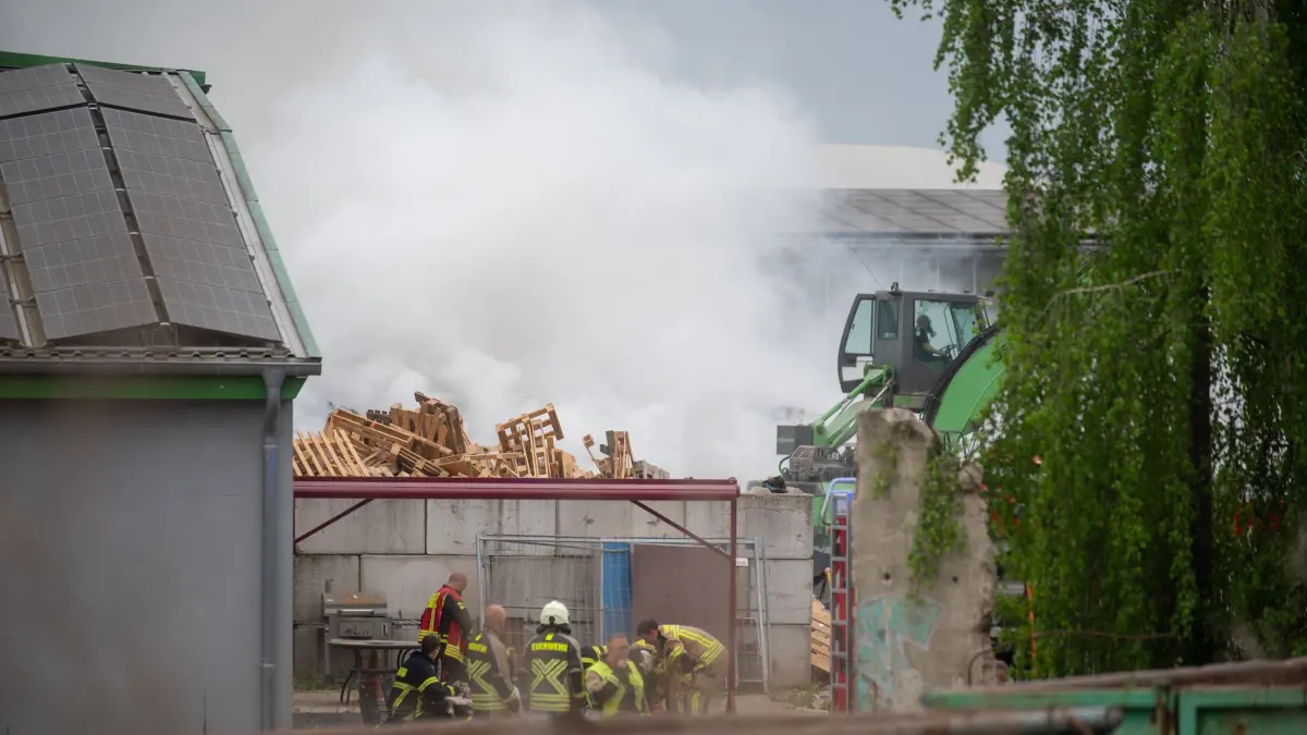 Mit einem Bagger wird der Sperrmüll auseinandergezogen, Feuerwehrkräfte löschen die Flammen.