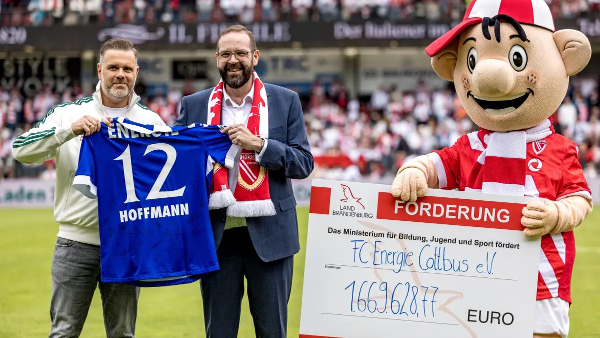 Energie Cottbus - SV Wehen Wiesbaden: 09.05.2026, Brandenburg, Cottbus: Fußball, Männer: 3. Liga, Energie Cottbus - SV Wehen Wiesbaden, 37. Spieltag, LEAG Energie Stadion. Sebastian Lemke (l-r,), Präsident des FC Energie Cottbus, Gordon Hoffmann (CDU), Minister für Bildung, Jugend und Sport des Landes Brandenburg, und Lauzi, Maskottchen, stehen bei der Übergabe eines Trikots an den Minister und eines Förderschecks an den Verein auf dem Spielfeld. Foto: Frank Hammerschmidt/dpa +++ dpa-Bildfunk +++