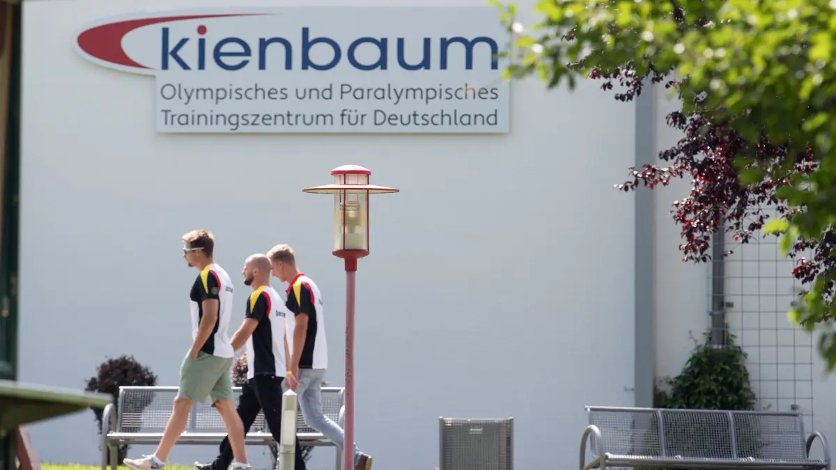 Drei Sportler gehen am 18.07.2017 im Bundesleistungszentrum in Kienbaum (Brandenburg) unter einem Schild mit der Aufschrift «Kienbaum Olympisches und Paralympisches Trainingszentrum für Deutschland». Am Nachmittag soll das Zentrum offiziell zum Olympischen und Paralympischen Trainingszentrum für Deutschland umbenannt und damit aufgewertet werden. Foto: Soeren Stache/dpa ++ +++ dpa-Bildfunk +++