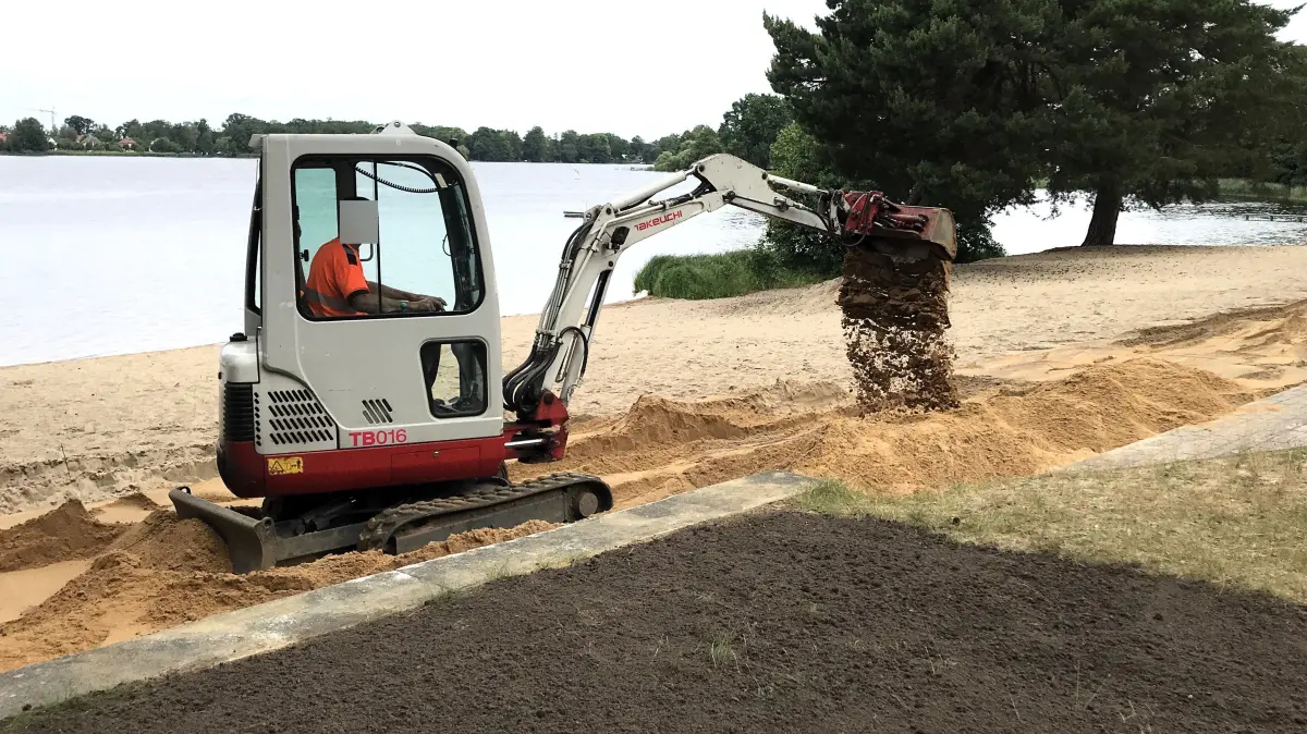 Müllrose. Freibad Großer Müllroser See. Nach dem Fund von unzähligen rostigen Metallteilen im Sand, Teile eines Maschendrahtzauns, ist auf einem Teil des Strandes der Sand ausgetauscht worden. Bild mit kleinem Bagger: Die Fahrrinnen des Baggers werden geglättet. 12.06.2026 Foto: Frank Groneberg