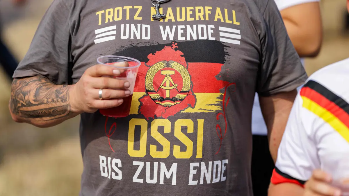 GERMANY-POLITICS-AFD-VOTE-ELECTION-PARTIES: A participant wears a shirt reading "despite the fall of the Wall and the political turnaround- Ossi to the end" during an election campaign event of the far-right Alternative for Germany (AfD) party's and a motorbike and moped ride in Jessnitz, eastern Germany on June 28, 2026. Fans of far-right Alternative for Germany party's top candidate for the regional elections in Saxony-Anhalt, Ulrich Siegmund joined him for a countryside ride on Simson-brand scooters, that have become a symbol of East German identity. Descendants of the Simson firm's namesake Jewish founders, who gave up the company to escape persecution in Nazi Germany, have denounced the AfD's appropriation of the brand name. (Photo by Jens SCHLÜTER / AFP)