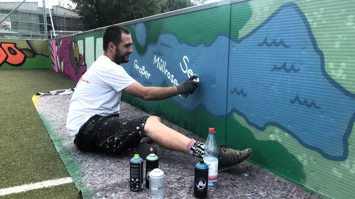 Müllrose. DFB-Minispielfeld in der Jahnstraße, Bolzplatz. Graffiti-Workshop Neugestaltung Bande. Oliver Bock, Künstlerkollektiv 153Design, stellt das Bild mit dem Großen Müllroser See, der um 90 Grad "gekippt" wurde, fertig. 16.07.2026 Foto: Frank Groneberg