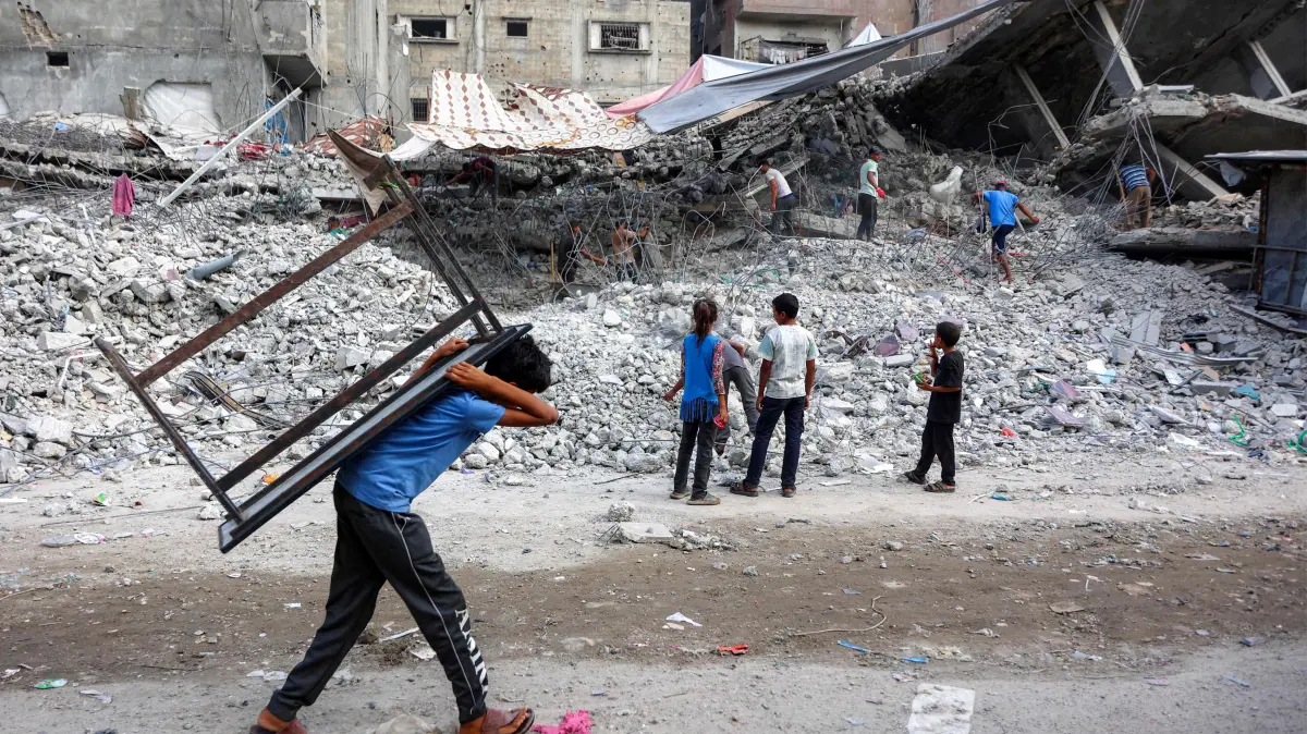 PALESTINIAN-ISRAEL-CONFLICT-GAZA-RECYCLING-RECONSTRUCTION: A boy walks carrying a table on his back past salvage workers scrapping the rubble of a building that was destroyed during Israel's war against Hamas in Gaza, to use the recycled remains in the future, in Gaza City on August 16, 2026. The Palestinian Islamist group Hamas had said on August 15 that it asked the US-backed Board of Peace to "compel" Israel to implement its obligations under the first phase of the US-brokered ceasefire plan in Gaza and to approve the roadmap for its second phase, after its leader met with the US president's envoy Jared Kushner in Egypt. (Photo by Omar AL-QATTAA / AFP)