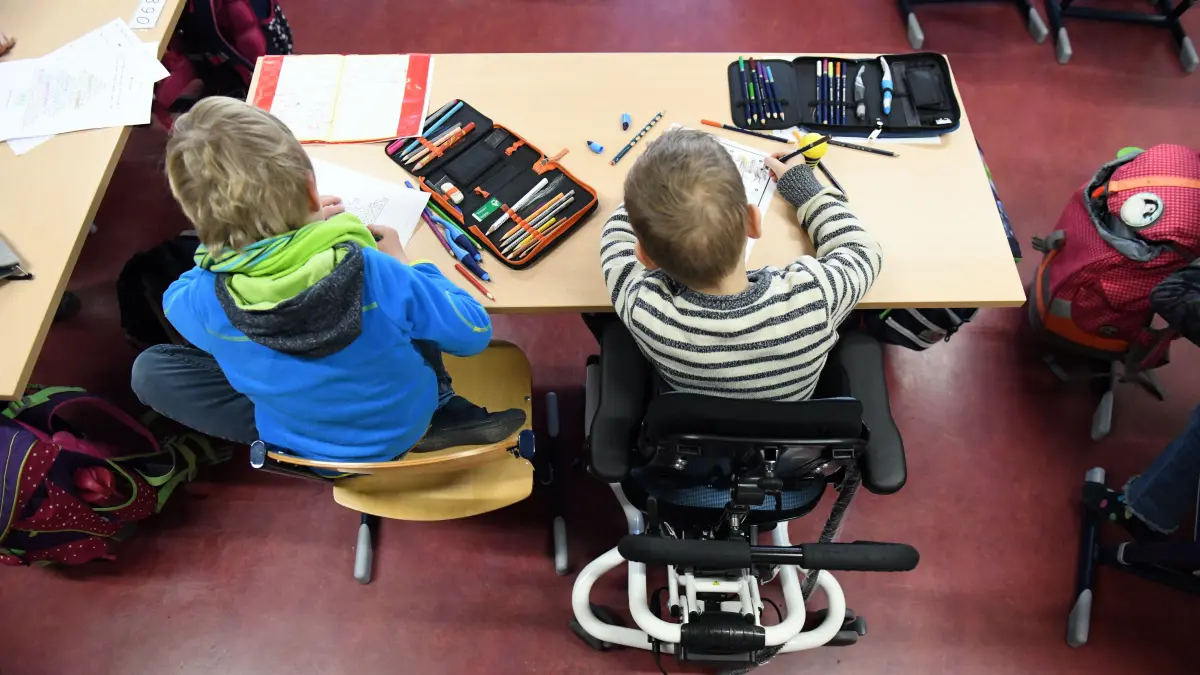 Ein Kind mit Behinderung sitzt an einer Grundschule in seinem Rollstuhl gemeinsam mit einem Erstklässler ohne Handicap in einem Klassenraum. Damit das „gemeinsame Lernen“ gut funktioniert, werden in einigen Fällen Schulbegleiter eingesetzt. Doch die Kosten dafür können die Kommunen kaum noch stemmen.