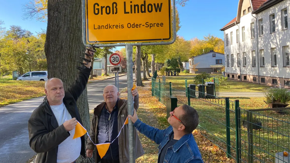 Kämpfen weiter um eine grundhafte Sanierung der Landesstraße 373: die Bürgermeister René Weiner (Groß Lindow), Horst Siebke (Brieskow-Finkenheerd) und der langjährige Amtsdirektor Dirk Wesuls (von links). Sie befestigen an zwei Ortseingangsschildern jeweils eine kaputte Autofeder und eine gelbe Girlande.
